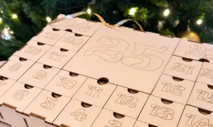 Calendario dell'avvento fatto a mano in legno, con numeri incisi e piccoli cassetti, sullo sfondo di un albero di Natale illuminato.