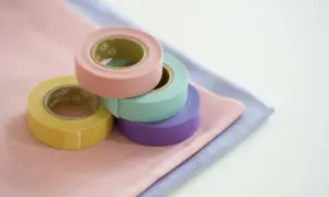 Rotoli di nastro adesivo per tessuti colorati (giallo, rosa, verde acqua, viola) su stoffe pastello. Utile per decorare o per un uso pratico su tessuti.