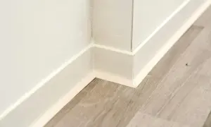 Angolo di una stanza con battiscopa bianco e pavimento in parquet effetto legno.