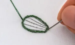 Dettaglio di schemi ricamo punto raso su tessuto bianco. Un ago con filo verde sta completando il contorno di una foglia.