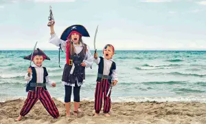 Tre bambini in costume da pirata fai da te, con spade e pistole giocattolo, esultano sulla spiaggia con il mare sullo sfondo.