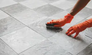 Mani con guanti arancioni che puliscono le fughe delle piastrelle con una spazzola, creando schiuma.