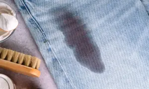 Un jeans con macchie di grasso scure. Accanto, una spazzola e una ciotola con bicarbonato, ingredienti per rimuovere le macchie.