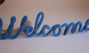 Lettera "Welcome" in filato blu, creata con un attrezzo per tricotin, appoggiata su un pezzo di legno.