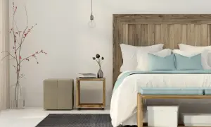 Un letto feng shui con testiera in legno grezzo, cuscini azzurri e bianchi, un vaso con rami rossi e un pouf beige.