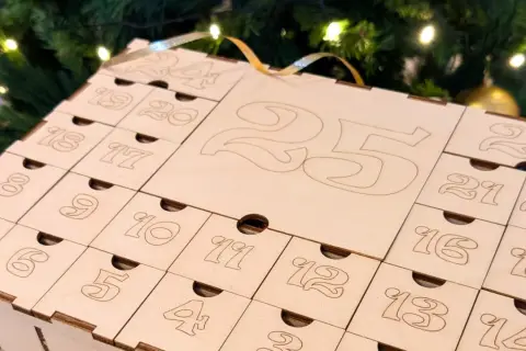 Calendario dell'avvento fatto a mano in legno, con numeri incisi e piccoli cassetti, sullo sfondo di un albero di Natale illuminato.