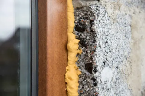 Schiuma espansa gialla sigilla i buchi tra un telaio di finestra marrone e un muro grezzo di cemento.