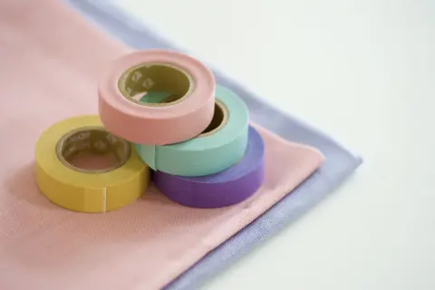 Rotoli di nastro adesivo per tessuti colorati (giallo, rosa, verde acqua, viola) su stoffe pastello. Utile per decorare o per un uso pratico su tessuti.