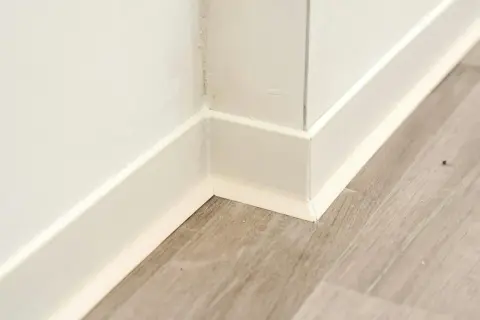 Angolo di una stanza con battiscopa bianco e pavimento in parquet effetto legno.