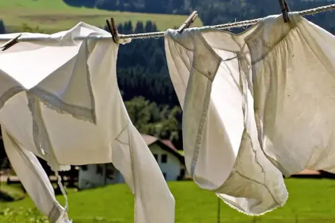 Capi ingialliti dal sole ondeggiano al vento su uno stendibiancheria, con un paesaggio verdeggiante e una casa sullo sfondo.