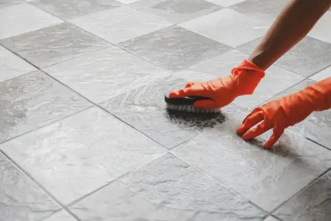Mani con guanti arancioni che puliscono le fughe delle piastrelle con una spazzola, creando schiuma.