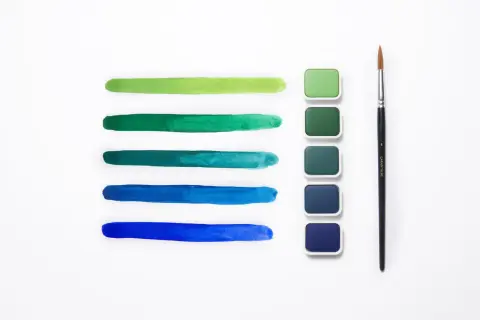 Pennellate verdi e blu mostrano la differenza tra colori acrilici e tempere, con pastiglie di colore e un pennello.