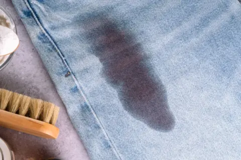 Un jeans con macchie di grasso scure. Accanto, una spazzola e una ciotola con bicarbonato, ingredienti per rimuovere le macchie.