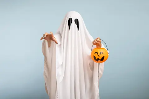 Bambino con un lenzuolo bianco e occhi neri, pronto per raccogliere dolcetti con la sua zucca arancione. Un'idea per costumi halloween fai da te bambini.