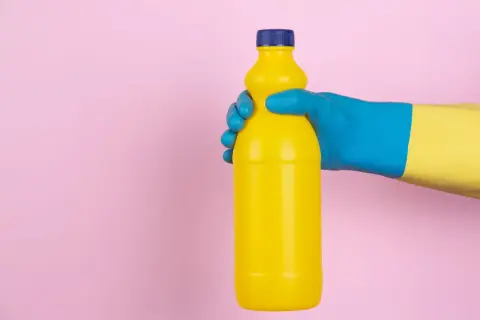 Mano con guanto blu e giallo tiene una bottiglia gialla. Attenzione, l'ammoniaca macchia i vestiti!