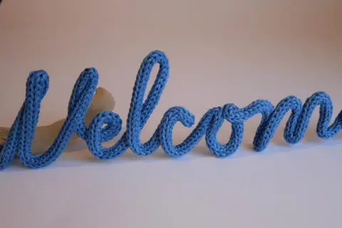 Lettera "Welcome" in filato blu, creata con un attrezzo per tricotin, appoggiata su un pezzo di legno.