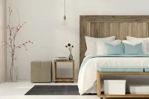 Un letto feng shui con testiera in legno grezzo, cuscini azzurri e bianchi, un vaso con rami rossi e un pouf beige.
