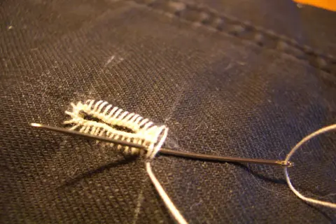 Un ago con filo bianco crea un punto occhiello a mano su tessuto scuro, un dettaglio di sartoria meticoloso.
