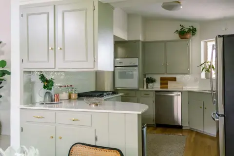 Cucina con mobili fai da te color salvia, piano bianco, elettrodomestici in acciaio e piante verdi.