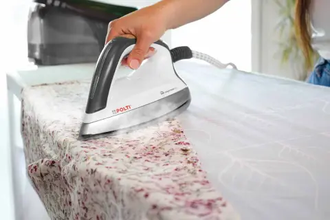 Mano che usa un ferro da stiro Polti per stirare una camicia a fiori. Il vapore aiuta a stirare la pelle del tessuto, rendendolo liscio.