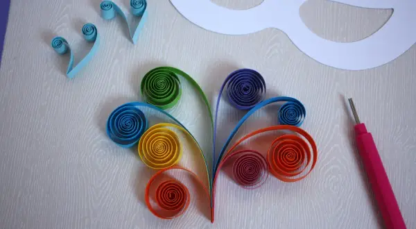 Creazione di una maschera carnevale fai da te con strisce di carta colorata arrotolate e uno strumento per quilling.