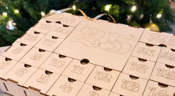 Calendario dell'avvento fatto a mano in legno, con numeri incisi e piccoli cassetti, sullo sfondo di un albero di Natale illuminato.