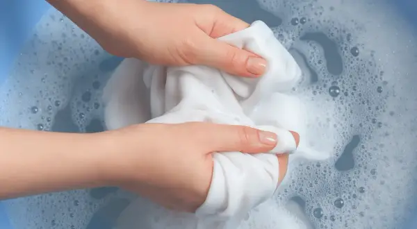 Mani che lavano un tessuto bianco in acqua saponata, creando schiuma e bollicine. L'acqua ossigenata sui tessuti aiuta a sbiancare.