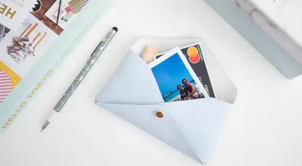 Pochette fai da te azzurra, con foto di coppia, carta di credito e banconota. Accanto, una penna e un libro.
