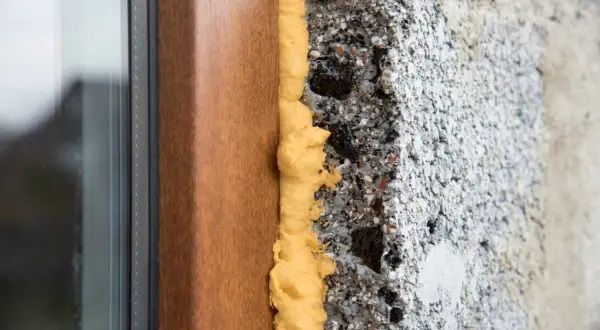 Schiuma espansa gialla sigilla i buchi tra un telaio di finestra marrone e un muro grezzo di cemento.