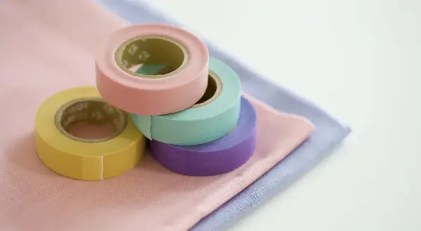 Rotoli di nastro adesivo per tessuti colorati (giallo, rosa, verde acqua, viola) su stoffe pastello. Utile per decorare o per un uso pratico su tessuti.