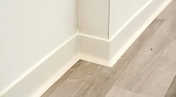 Angolo di una stanza con battiscopa bianco e pavimento in parquet effetto legno.