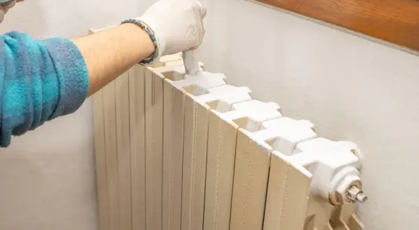 Mano con guanto bianco che applica vernici per termosifoni con un pennello. Il radiatore è beige e la vernice è bianca.