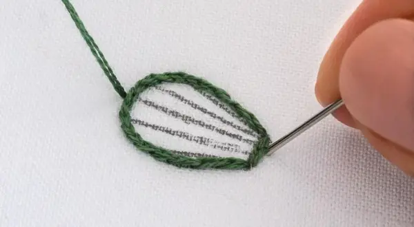 Dettaglio di schemi ricamo punto raso su tessuto bianco. Un ago con filo verde sta completando il contorno di una foglia.