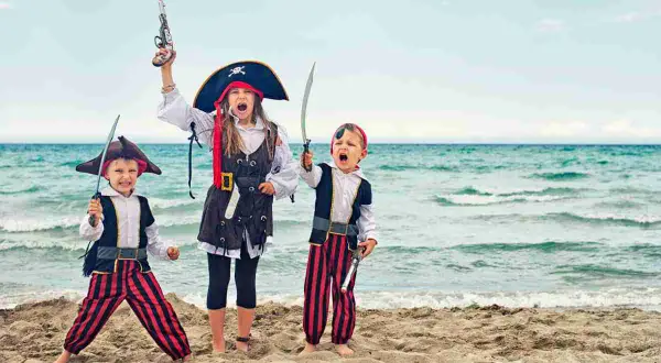 Tre bambini in costume da pirata fai da te, con spade e pistole giocattolo, esultano sulla spiaggia con il mare sullo sfondo.