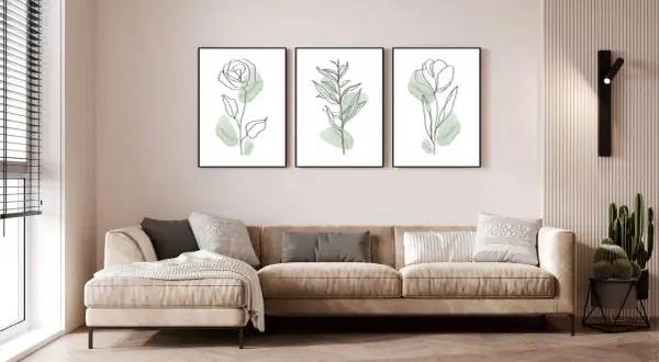 Divano beige in un soggiorno al piano terra poco luminoso, con tre quadri botanici minimalisti e una lampada da parete.