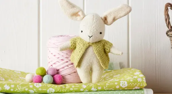 Coniglietto di stoffa con giacca verde, accanto a gomitolo rosa e palline colorate. Un cartamodello coniglio stoffa per creare il tuo amico.