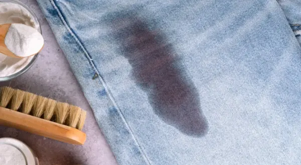 Un jeans con macchie di grasso scure. Accanto, una spazzola e una ciotola con bicarbonato, ingredienti per rimuovere le macchie.