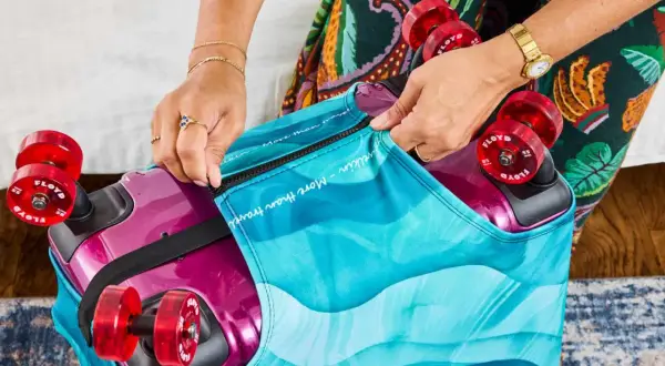 Hands zipping a colorful, wave-patterned cover onto a magenta suitcase with red wheels.