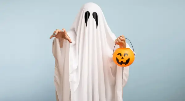 Bambino con un lenzuolo bianco e occhi neri, pronto per raccogliere dolcetti con la sua zucca arancione. Un'idea per costumi halloween fai da te bambini.