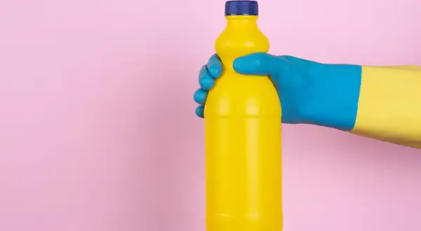 Mano con guanto blu e giallo tiene una bottiglia gialla. Attenzione, l'ammoniaca macchia i vestiti!