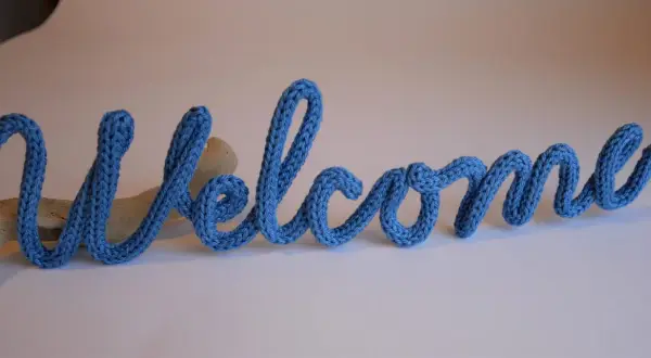 Lettera "Welcome" in filato blu, creata con un attrezzo per tricotin, appoggiata su un pezzo di legno.