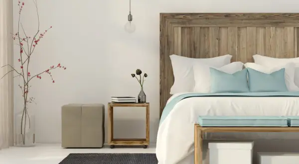 Un letto feng shui con testiera in legno grezzo, cuscini azzurri e bianchi, un vaso con rami rossi e un pouf beige.