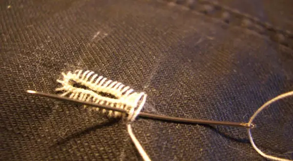 Un ago con filo bianco crea un punto occhiello a mano su tessuto scuro, un dettaglio di sartoria meticoloso.