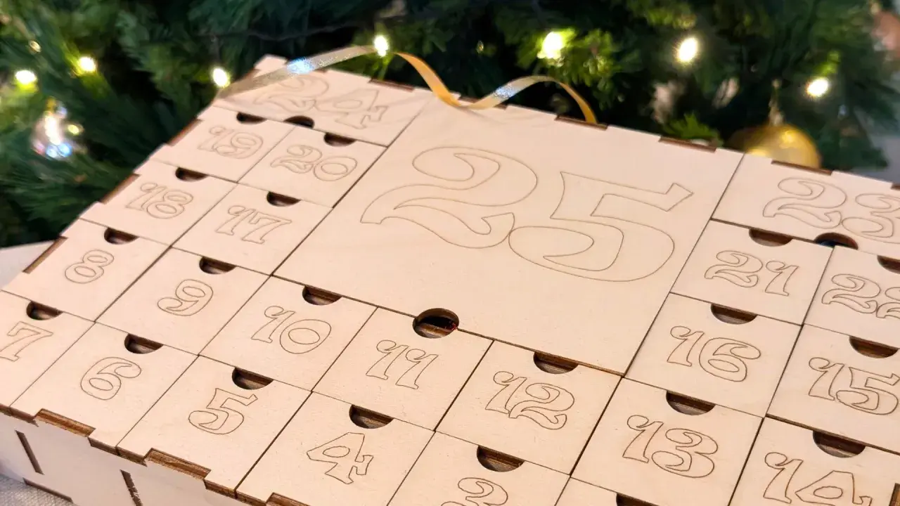 Calendario dell'avvento fatto a mano in legno, con numeri incisi e piccoli cassetti, sullo sfondo di un albero di Natale illuminato.