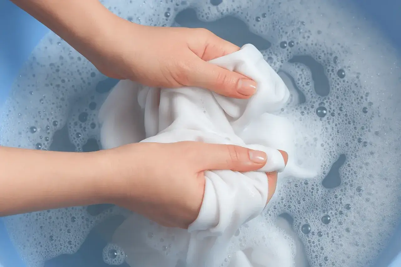 Mani che lavano un tessuto bianco in acqua saponata, creando schiuma e bollicine. L'acqua ossigenata sui tessuti aiuta a sbiancare.