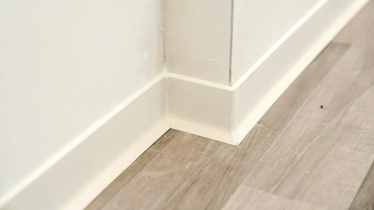 Angolo di una stanza con battiscopa bianco e pavimento in parquet effetto legno.