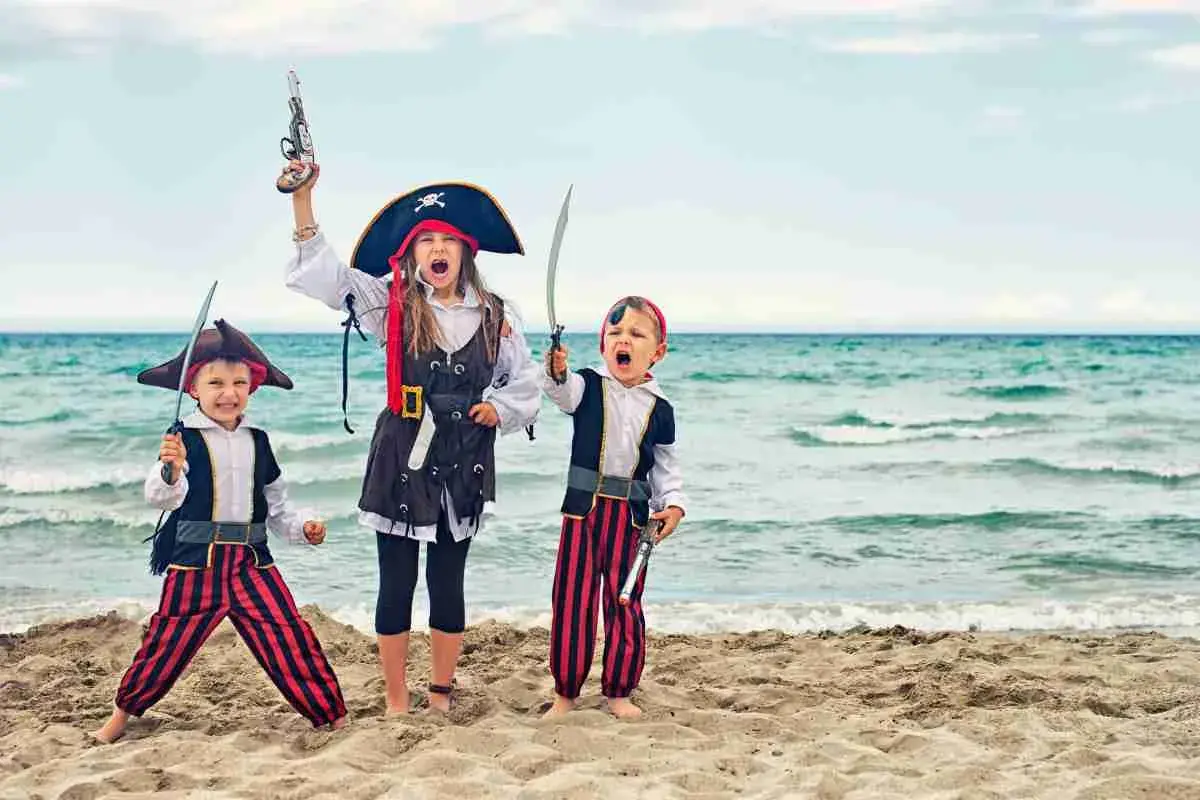 Tre bambini in costume da pirata fai da te, con spade e pistole giocattolo, esultano sulla spiaggia con il mare sullo sfondo.