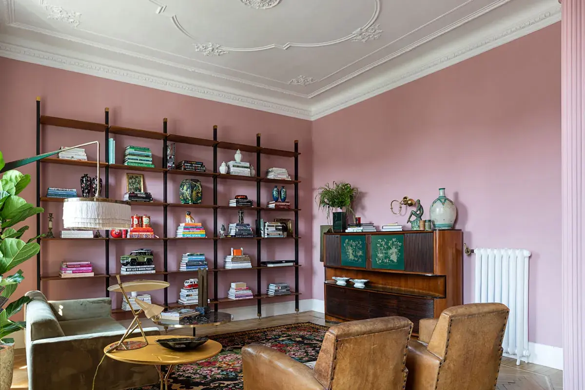 Soggiorno accogliente con **caldi colori pareti** rosa cipria, libreria a giorno piena di libri e oggetti, divano verde, poltrone in pelle e mobile vintage.