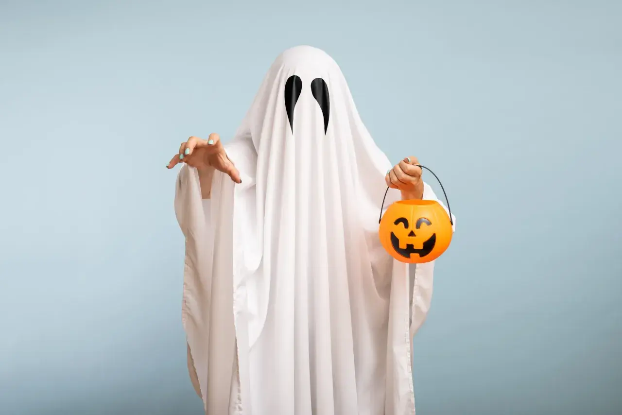 Bambino con un lenzuolo bianco e occhi neri, pronto per raccogliere dolcetti con la sua zucca arancione. Un'idea per costumi halloween fai da te bambini.