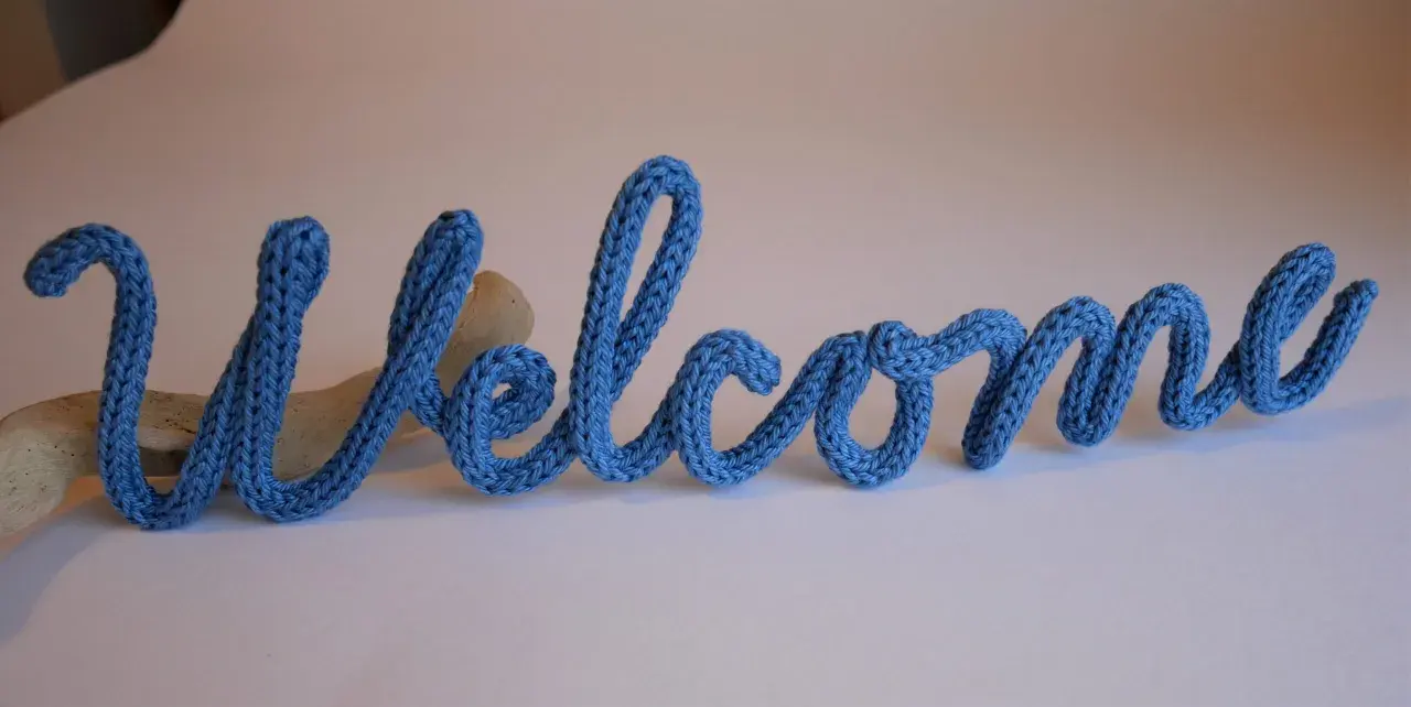 Lettera "Welcome" in filato blu, creata con un attrezzo per tricotin, appoggiata su un pezzo di legno.